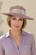A woman wearing a wide-brimmed mauve hat with a patterned band, a mauve button-up shirt, and white pants, standing against a textured beige wall.