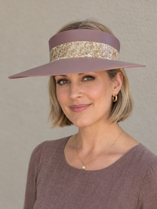 A woman wearing a dusty mauve wide-brimmed sun visor with a gold and white patterned band, paired with a matching mauve long-sleeve top, set against a neutral beige wall.