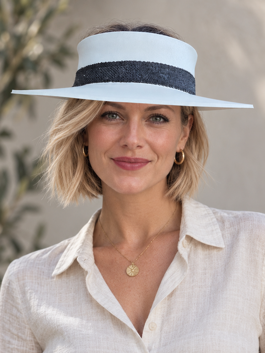 A woman wearing a wide-brimmed white sun visor with a dark navy band, a light beige linen button-down shirt, and a woven belt, set against a soft-focus outdoor background.