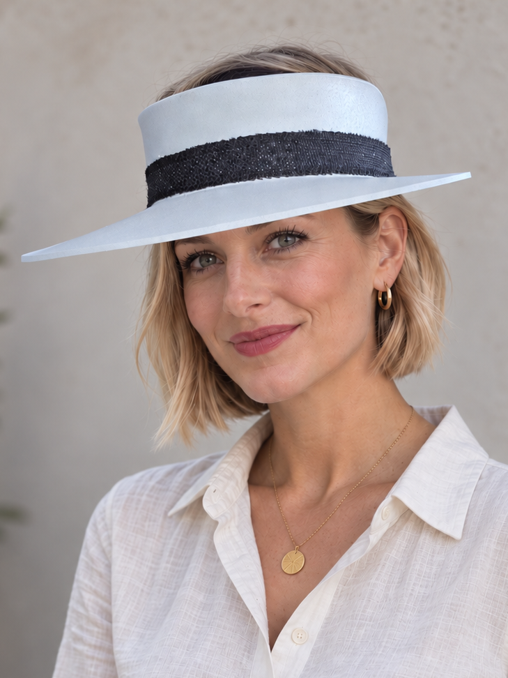 A woman wearing a white wide-brimmed sun hat with a black band and a cream-colored linen shirt, standing against a textured beige wall.