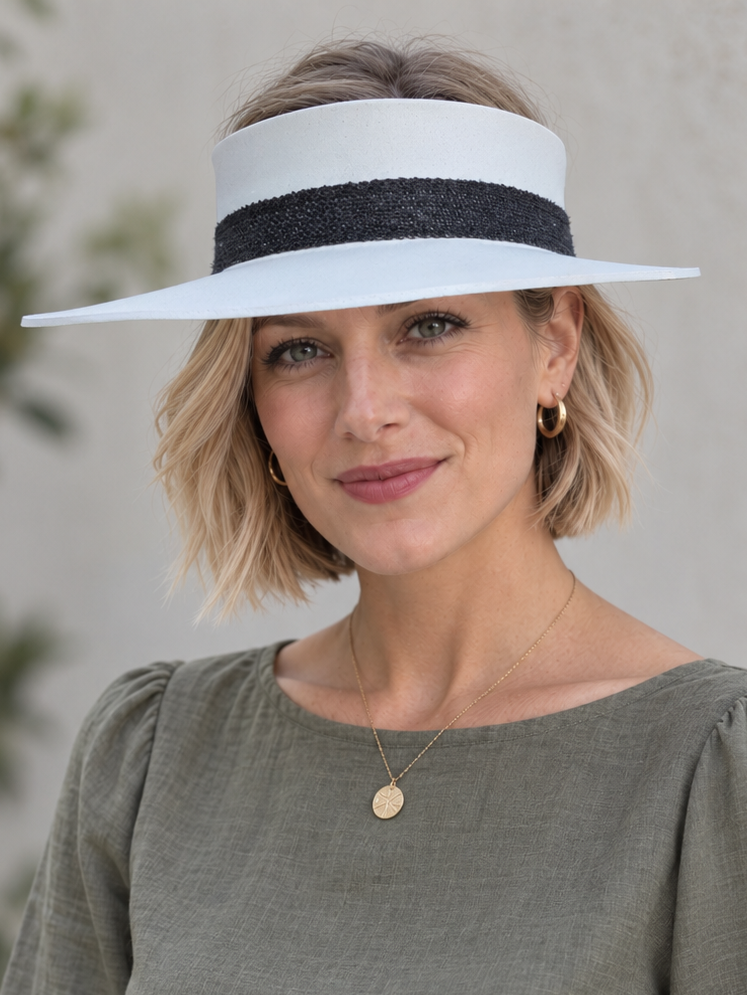 A woman wearing a wide-brimmed white sun visor with a black textured band, paired with a sage green long-sleeve blouse and white pants, photographed against a soft-focus outdoor background.