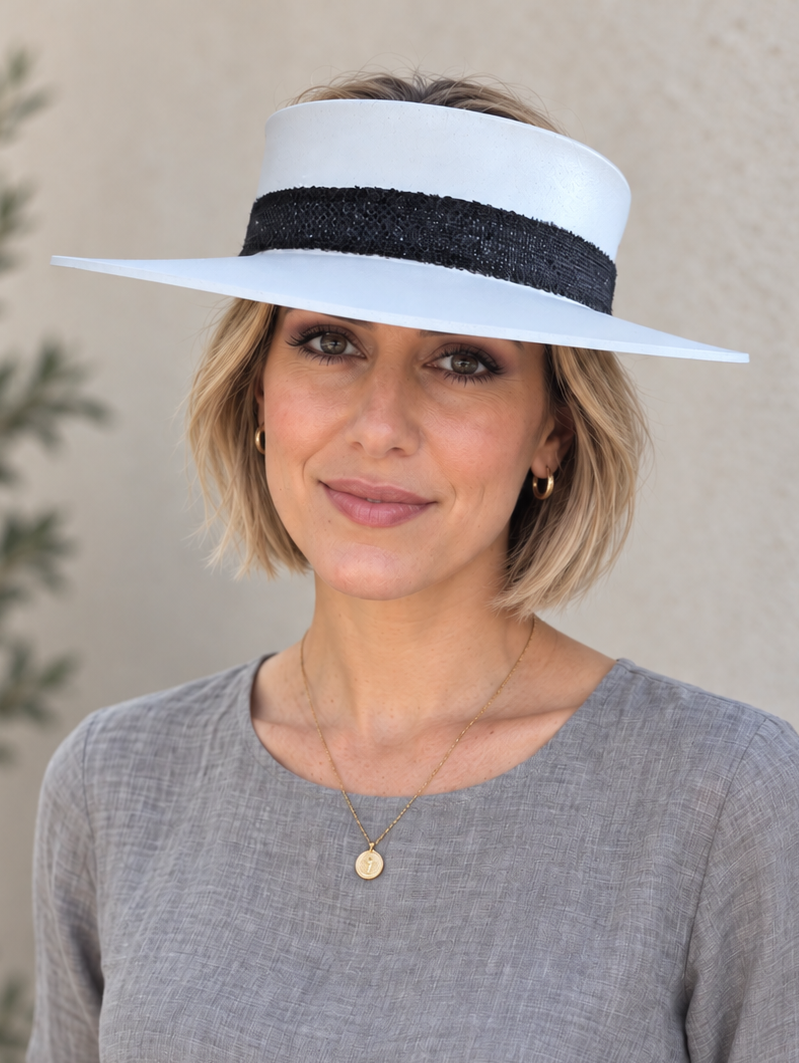 A woman wearing a white wide-brimmed sun visor hat with a black band, a gray linen long-sleeve top, and white pants, standing against a textured beige wall.