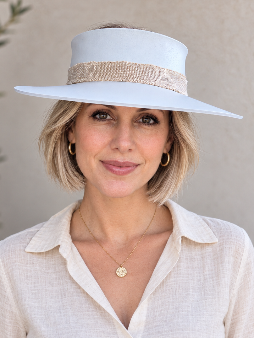 A woman wearing a wide-brimmed white sun hat with a textured beige band and a cream linen button-up shirt, posing against a neutral beige wall.