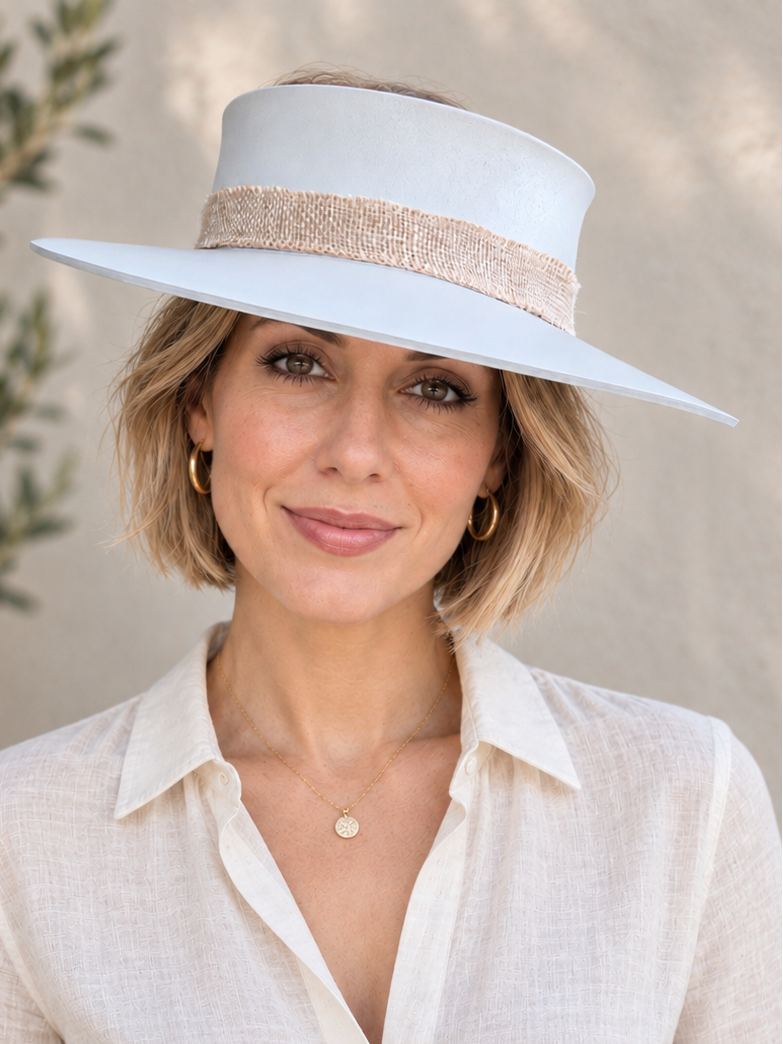 A woman wearing a wide-brimmed white sun hat with a beige textured band and a cream linen button-up shirt, posed against a neutral stucco wall with soft shadows.
