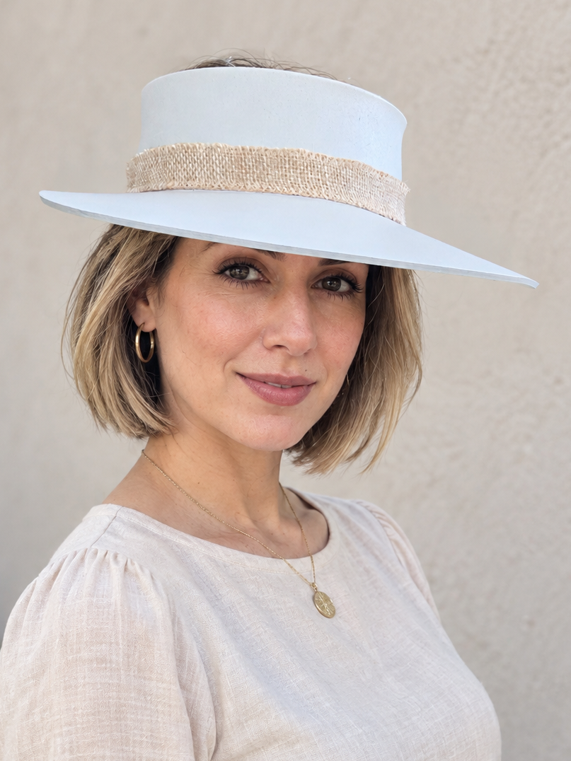 A woman wearing a white wide-brimmed sun hat with a textured beige band and a light beige linen blouse, standing against a textured beige stucco wall.