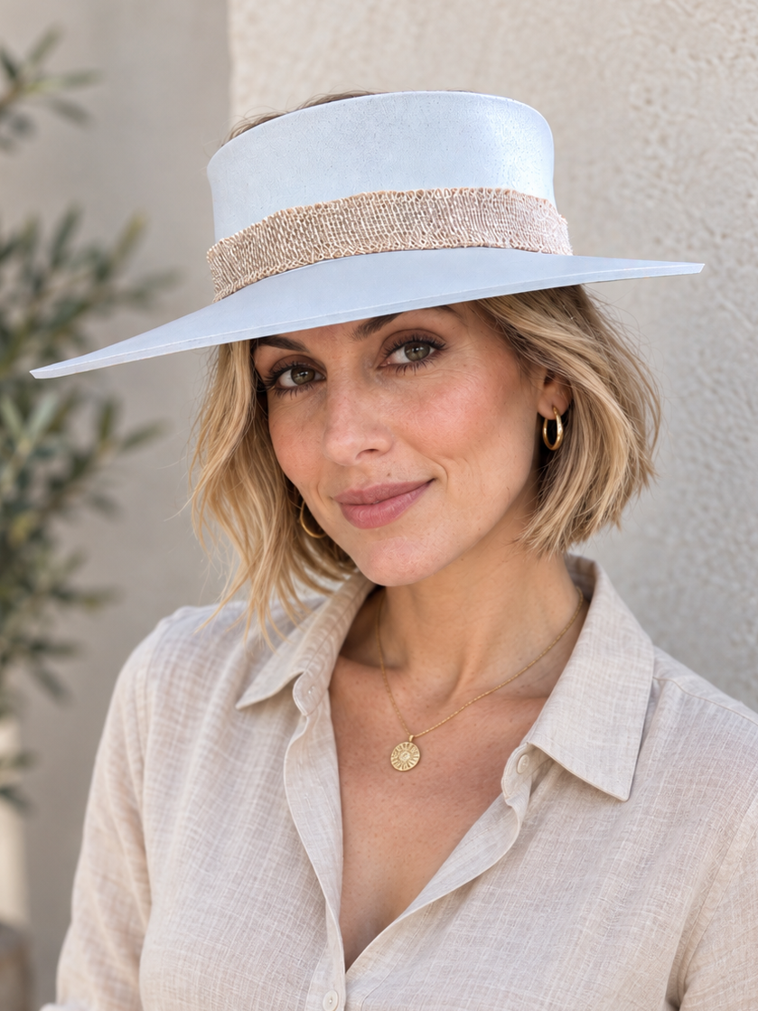 A woman wearing a wide-brimmed white sun hat with a beige woven band and a light beige linen button-up shirt, posing against a textured beige wall.