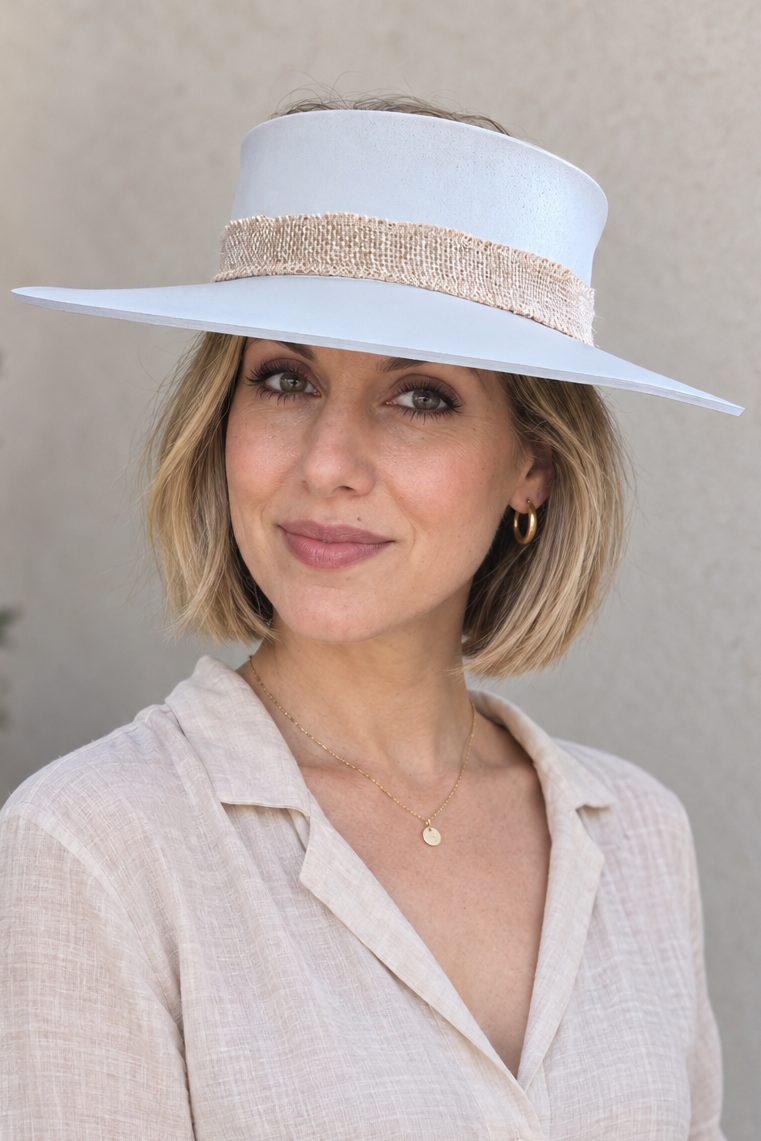 A woman wearing a wide-brimmed white sun hat with a beige textured band and a light beige linen button-up shirt, posed against a neutral stucco wall.