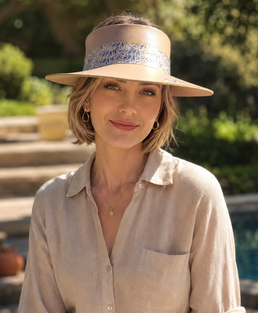 A woman wearing a beige wide-brim sun hat with a blue patterned band, a light beige linen button-up shirt, and white pants, sitting outdoors near a pool with a blurred garden background.