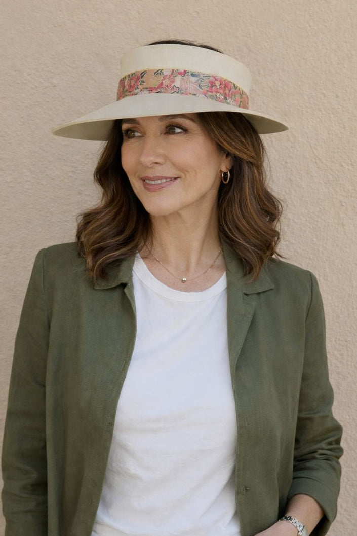 Woman wearing a green jacket, white shirt, and floral visor hat against a beige wall.