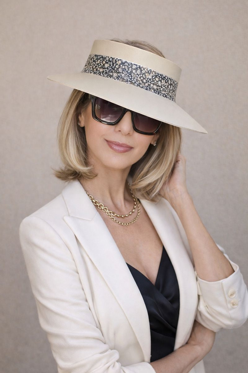 Woman wearing a white sun hat with a decorative band, sunglasses, and a white blazer against a neutral background.