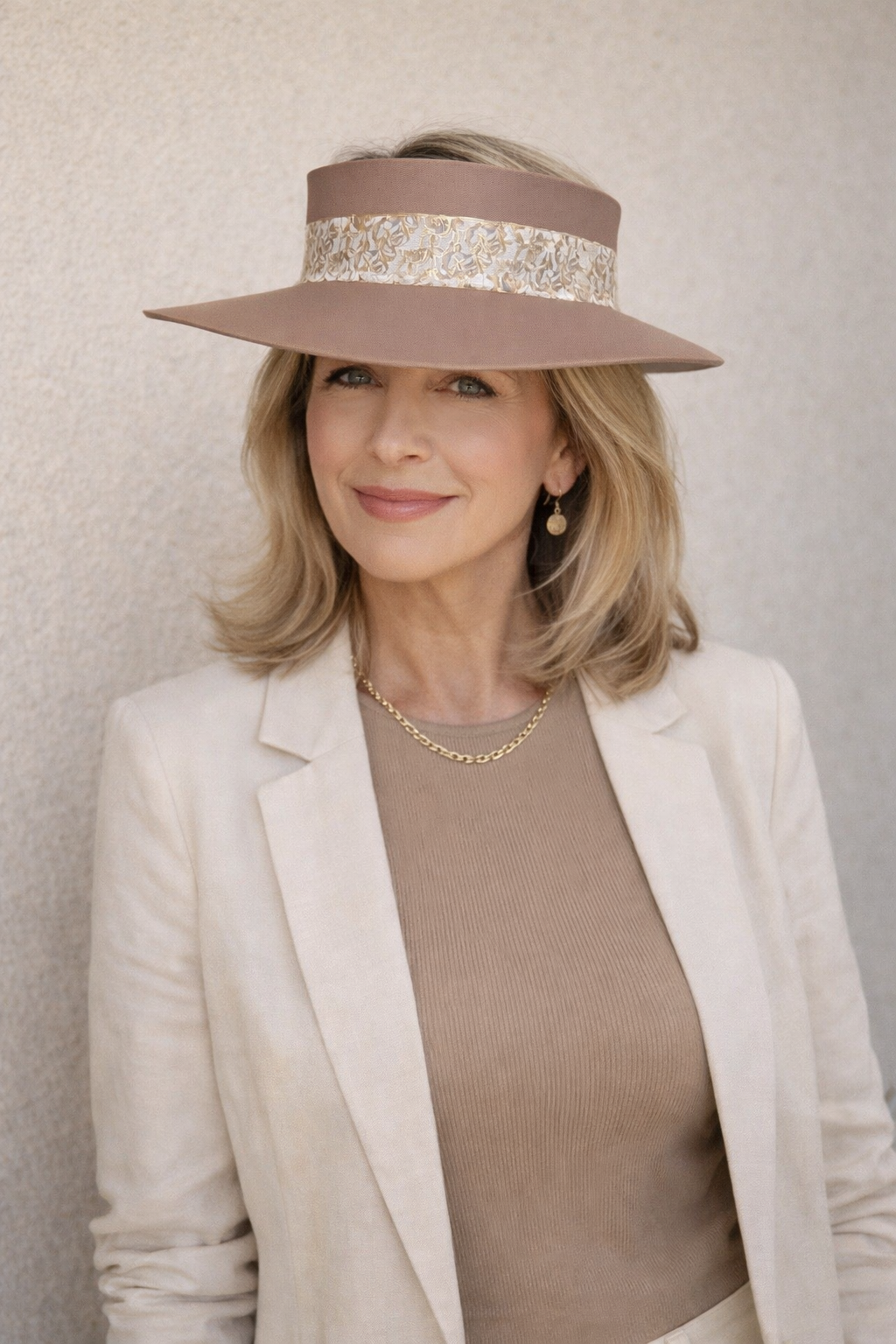Woman wearing a beige hat with a decorative band, beige blazer, and brown top against a neutral background.