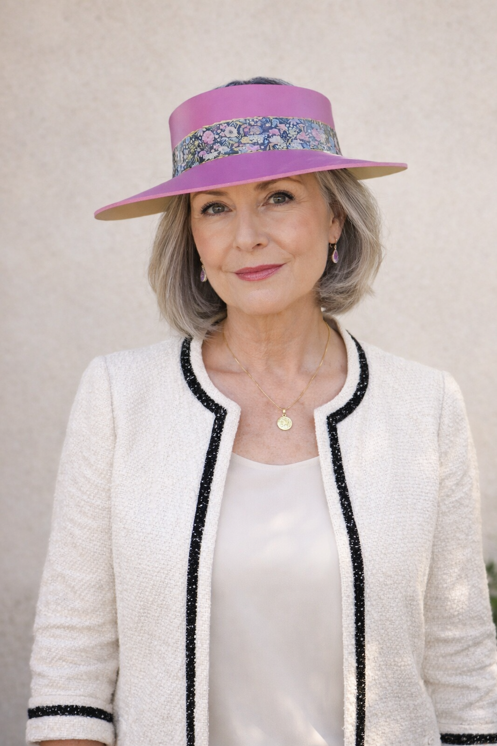 Woman wearing a white jacket with black trim and a pink hat with floral band against a plain background