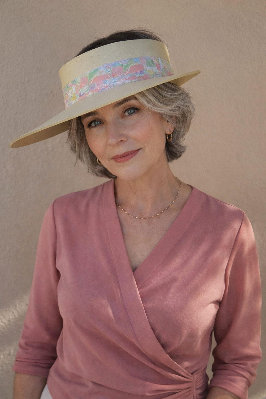 Woman wearing a wide-brimmed hat with floral band and a pink top against a beige wall.