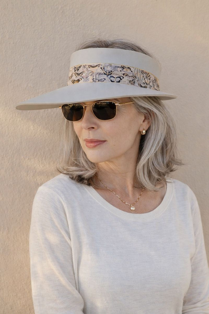 Woman wearing a wide-brimmed hat with floral band and sunglasses against a beige wall.