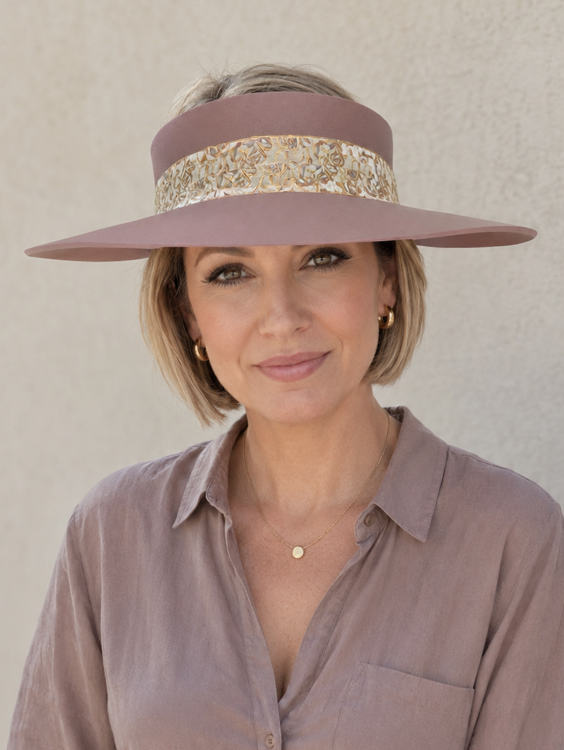 A woman wearing a dusty mauve wide-brim sun hat with a gold patterned band and a matching mauve button-down shirt, posing against a neutral beige wall.