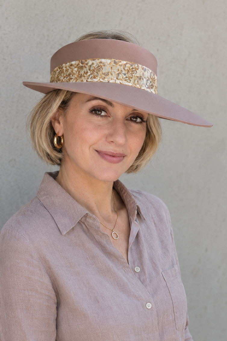 A woman wearing a dusty pink wide-brim sun visor hat with a gold sequin band, paired with a matching dusty pink linen button-down shirt and cream pants, standing against a textured gray wall.