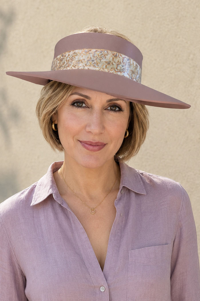 A woman wearing a wide-brimmed mauve hat with a patterned band, a mauve button-up shirt, and white pants, standing against a textured beige wall.