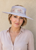 A woman wearing a wide-brimmed lavender sun hat with a floral band and a light pink linen button-up shirt, standing against a textured beige stucco wall.