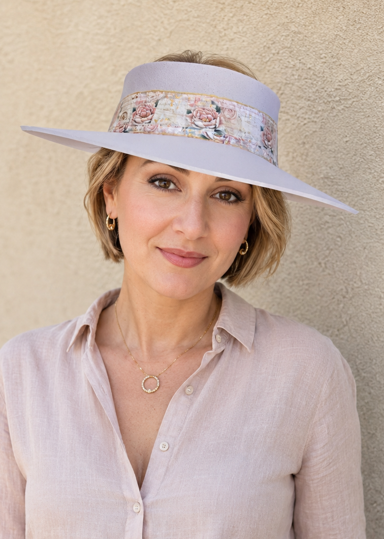 A woman wearing a wide-brimmed lavender sun hat with a floral band and a light pink linen button-up shirt, standing against a textured beige stucco wall.