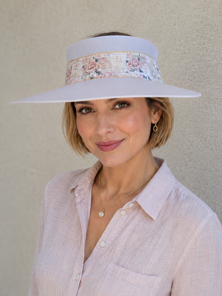 A woman wearing a wide-brimmed white sun visor with a pastel floral band and a light pink linen button-up shirt, standing against a textured beige stucco wall.