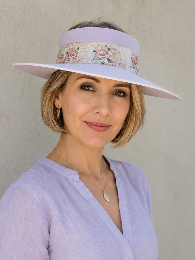 A woman wearing a lavender wide-brim sun visor hat with a floral band and a matching lavender long-sleeve top, standing against a textured beige wall.