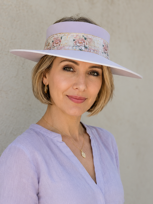 A woman wearing a lavender wide-brim sun visor hat with a floral band and a matching lavender long-sleeve top, standing against a textured beige wall.