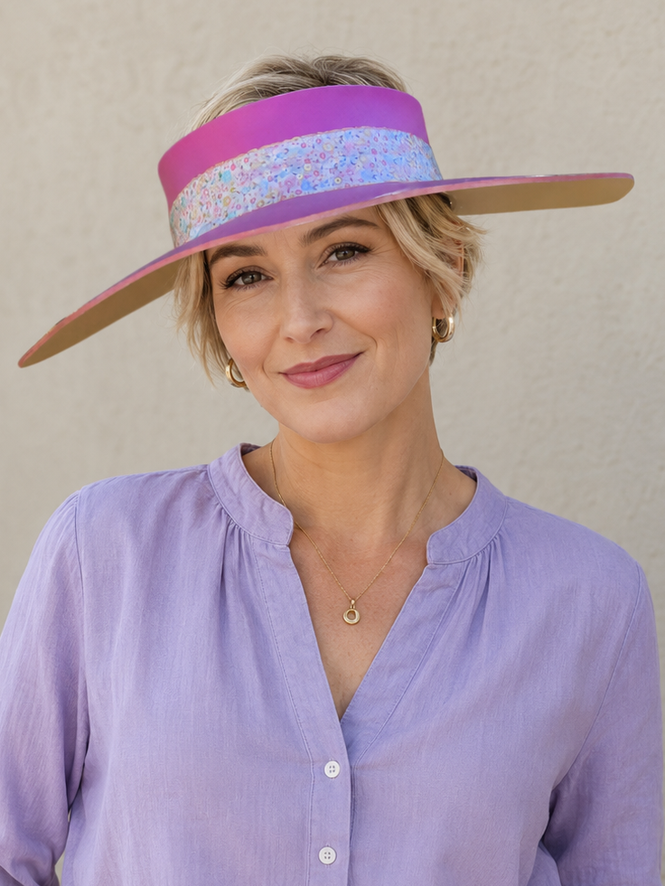 Woman wearing a wide-brimmed hat with a colorful band, a light purple shirt, and white pants against a beige background.