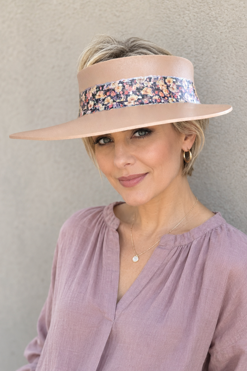 A woman wearing a wide-brimmed beige sun visor with a colorful floral pattern band, paired with a dusty purple blouse and white pants, posing against a textured gray wall.