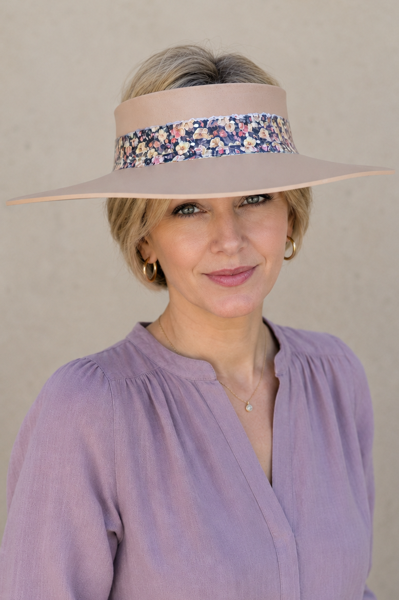 A woman wearing a wide-brimmed beige sun visor with a colorful floral pattern band, paired with a lavender long-sleeve blouse and white pants, set against a neutral beige background.