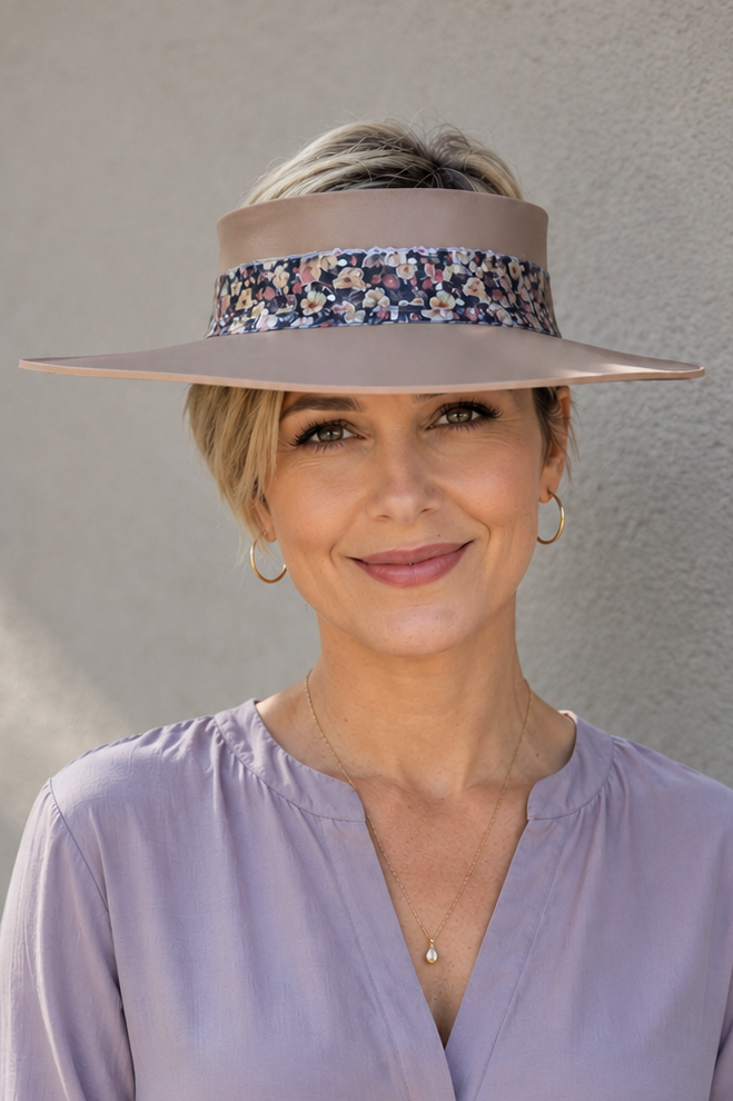 A woman wearing a wide-brimmed beige sun hat with a colorful floral pattern band, paired with a lavender blouse and white pants, standing against a textured gray wall.