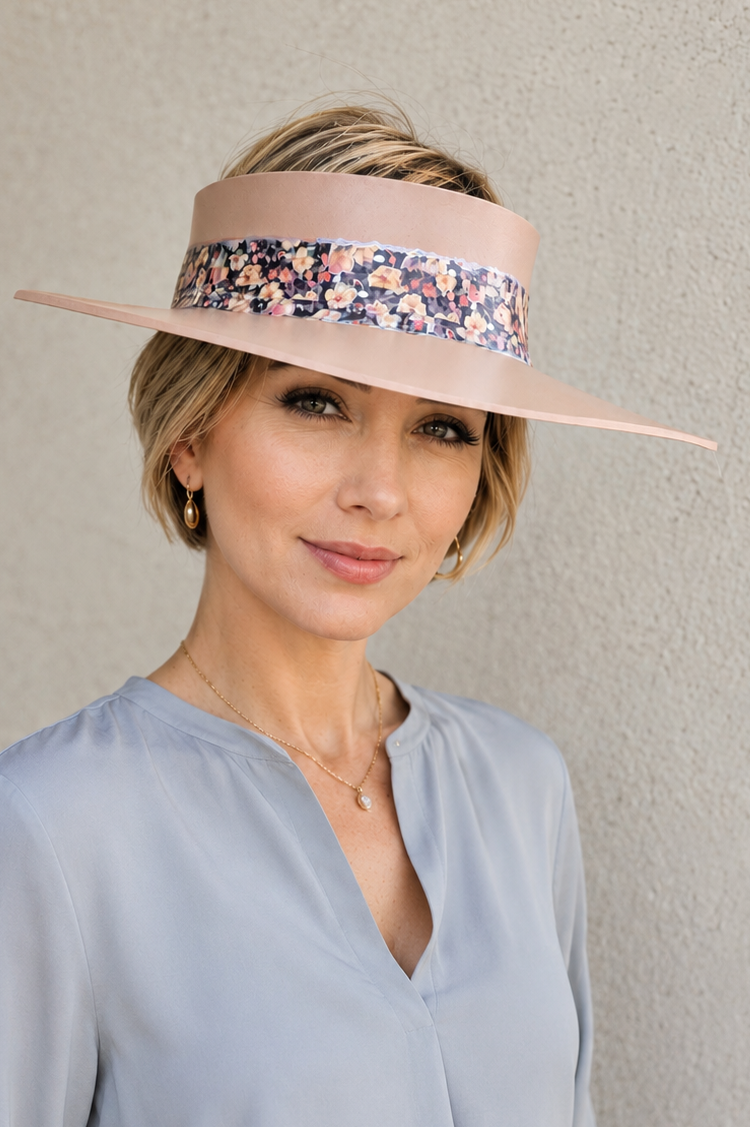 A woman wearing a light pink wide-brim sun hat with a floral band and a light blue blouse, standing against a textured gray wall.