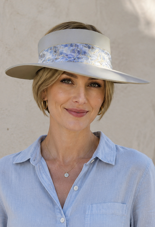 Woman wearing a light blue shirt and a wide-brimmed hat with floral band against a beige wall.