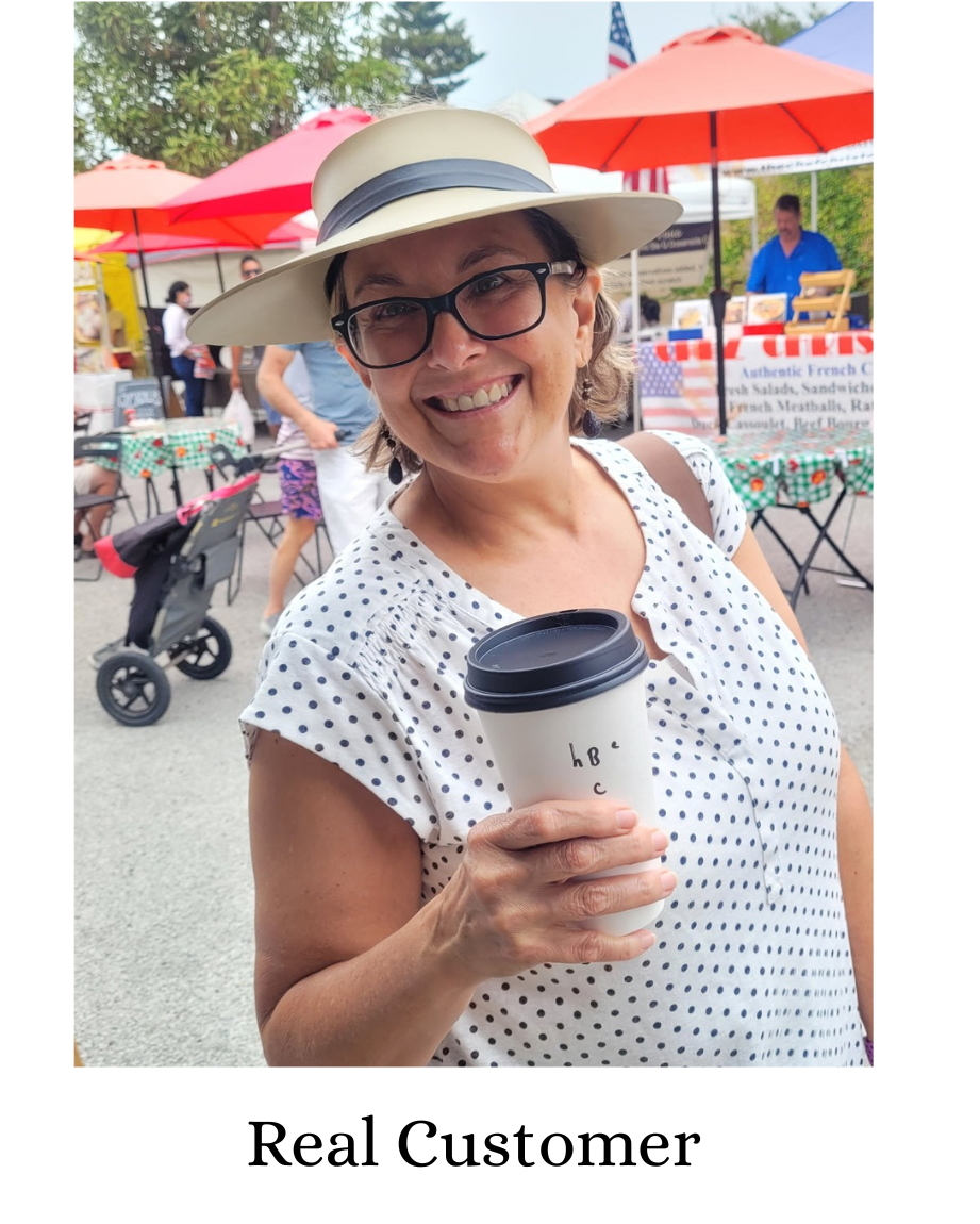 A smiling woman wearing a beige sun hat with a black band, black-rimmed glasses, and a white sleeveless top with small black polka dots holds a paper coffee cup. The photo is taken outdoors at what looks like a market or event, with red umbrellas and tables in the background.