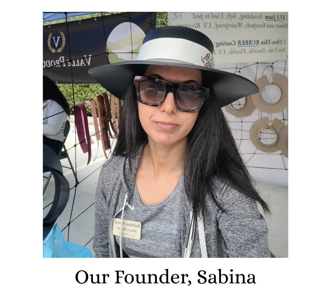 A woman wearing a black wide-brim sun hat with a white band, dark sunglasses, and a gray top, standing outdoors in front of a booth. Text below reads 'Our Founder, Sabina'.
