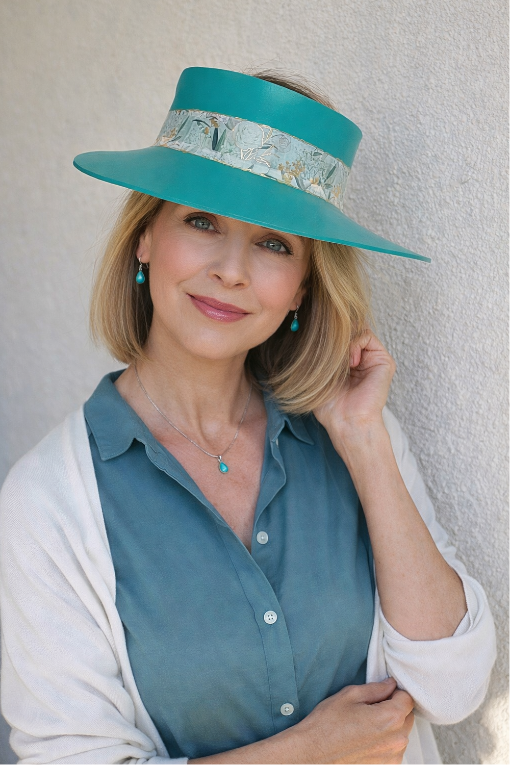 Woman wearing a teal hat with a white pattern, blue shirt, and white cardigan against a light background